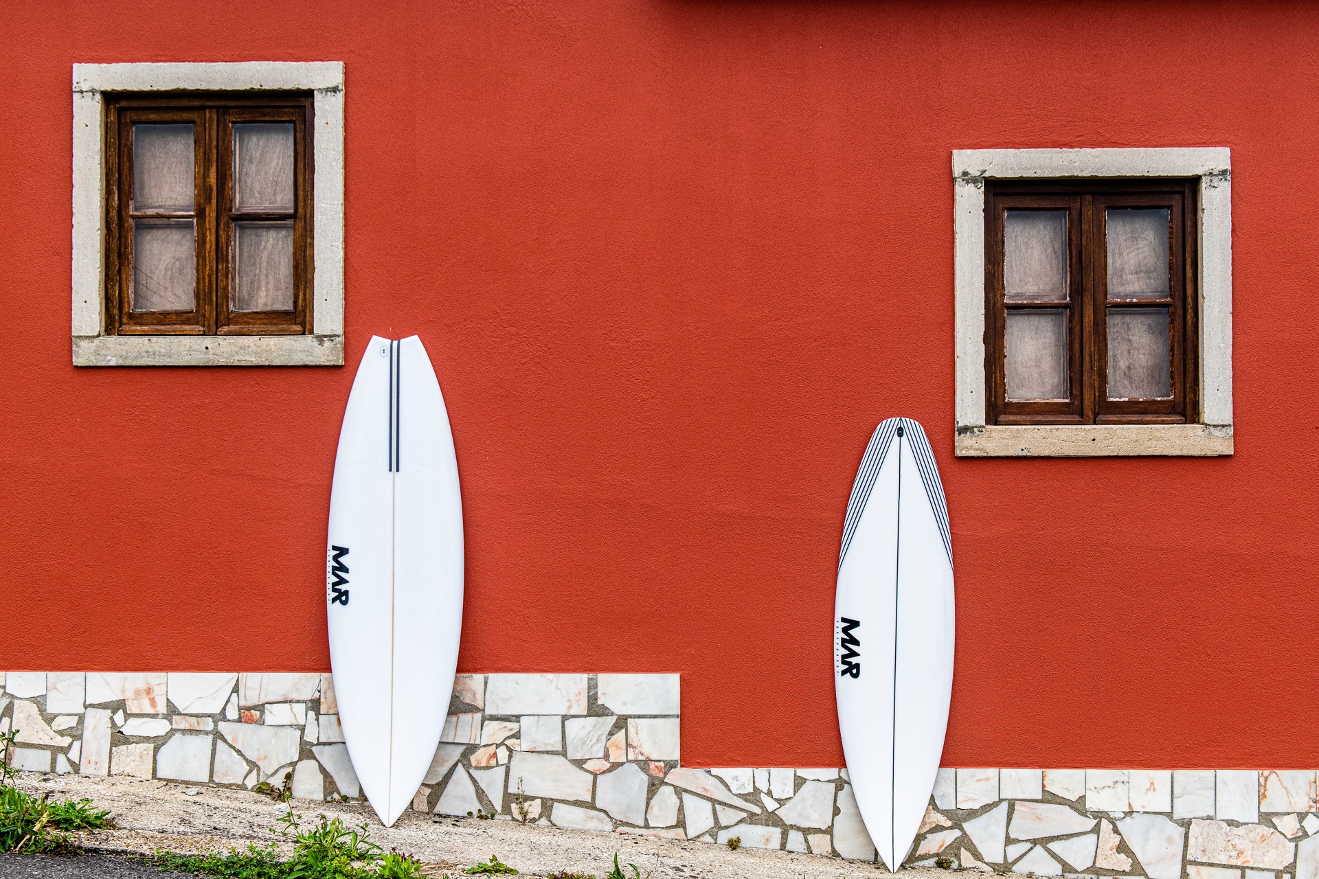 Two white MAR surfboards leaning against a textured red wall beneath wooden framed windows in an urban outdoor setting.