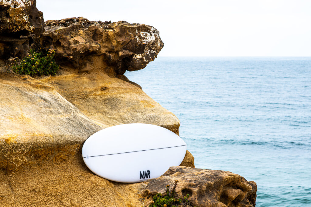 MAR Surfboards white surfboard resting on a rocky coastline overlooking the ocean.