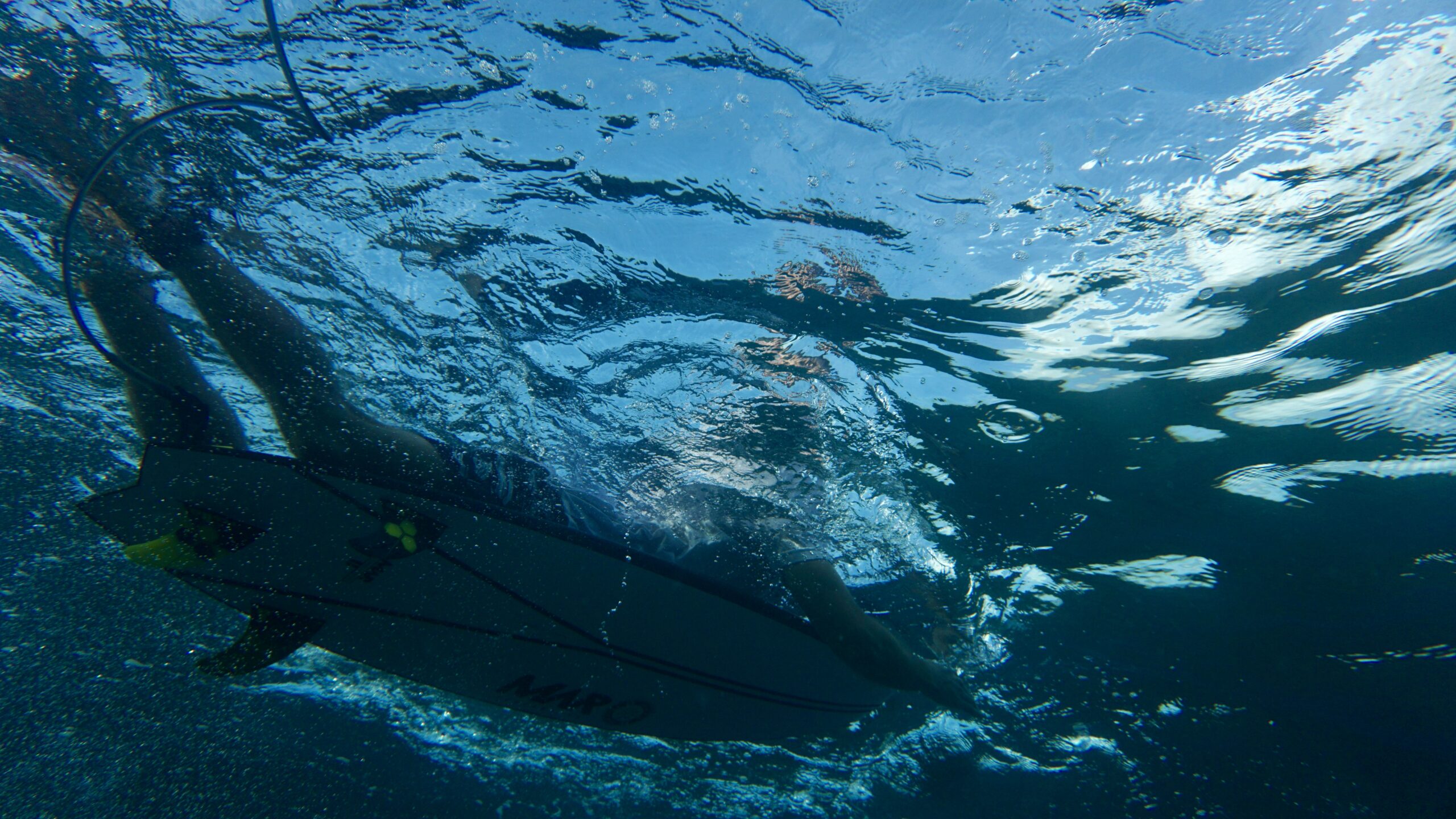 Underwater view of a surfer paddling a MAR Surfboards performance surfboard through clear ocean water.