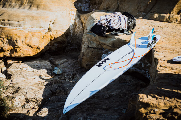 MAR Surfboards performance surfboard resting on rugged coastal rocks before a surf session.